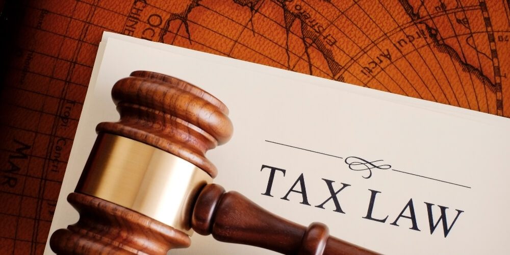 Wooden gavel on a document titled "Tax Law" atop a world map background. The image conveys a serious and authoritative tone.