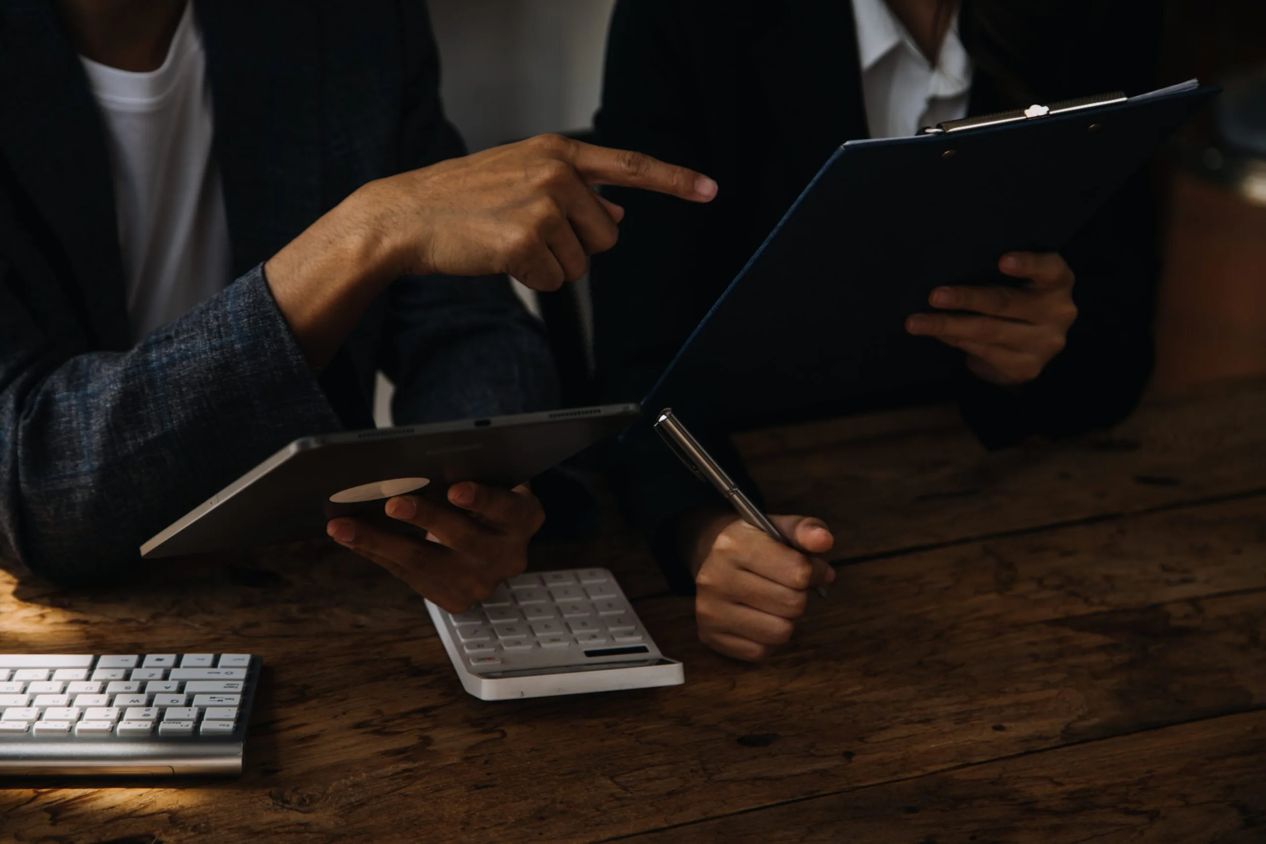 Two people in business attire work at a wooden desk with a tablet, a clipboard, a white calculator, and a keyboard, one pointing at the documents.