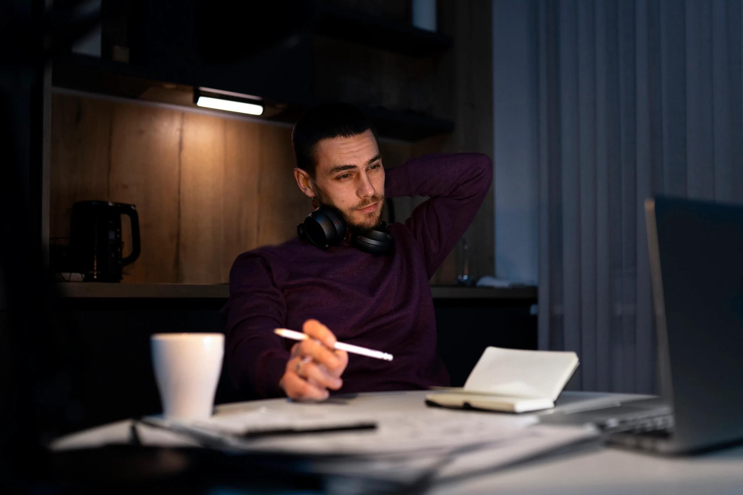 A man in a purple sweater with headphones around his neck sits at a desk, holding a pen near an open notebook while looking toward a laptop screen.