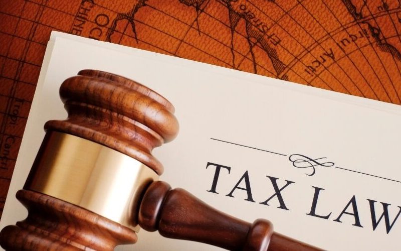 Wooden gavel on a document titled "Tax Law" atop a world map background. The image conveys a serious and authoritative tone.